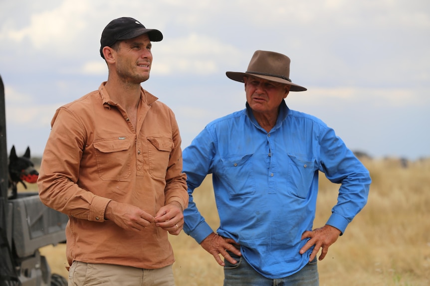 Two men stand outside in farming paddock, one wears a cap and another a hat.
