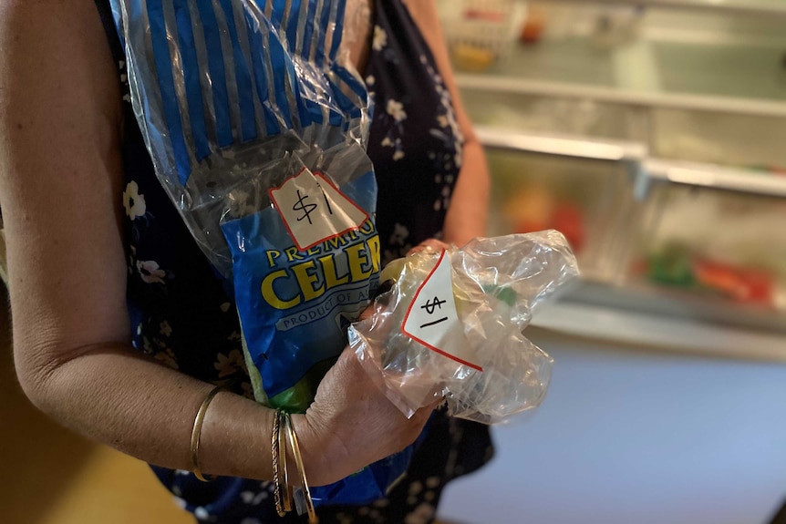 a woman in a blue top holds a plastic package of celery marked $1 and another plastic packaged marked $1.