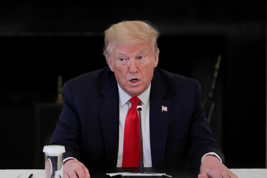 A man wearing a dark suit with a red tie puts his hands on a table near a glass of water