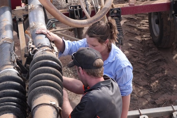 In this photo, taken form above, a man in a cap and woman in a blue short look attentively at a piece of farm machinery