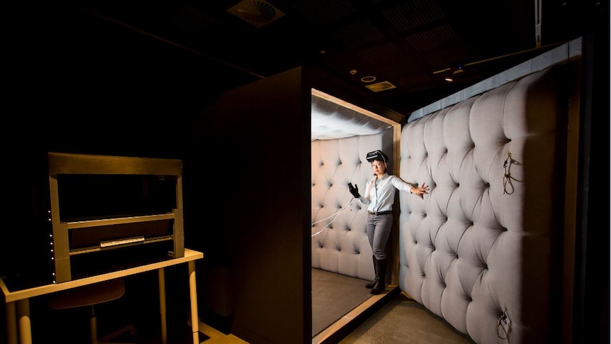 A woman with a VR headset stands at the entrance of a small room with padding on the interior walls