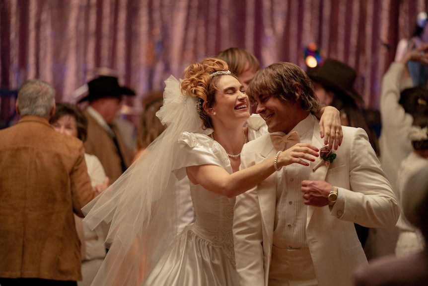 A film still of Lily James and Zac Efron as bride and groom, in 80s-style outfits. They're smiling brightly and embracing.