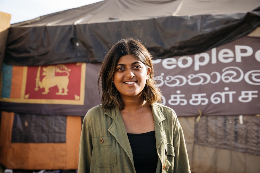 A young Sri Lankan woman in a green jacket smiles while standing in front of tents 