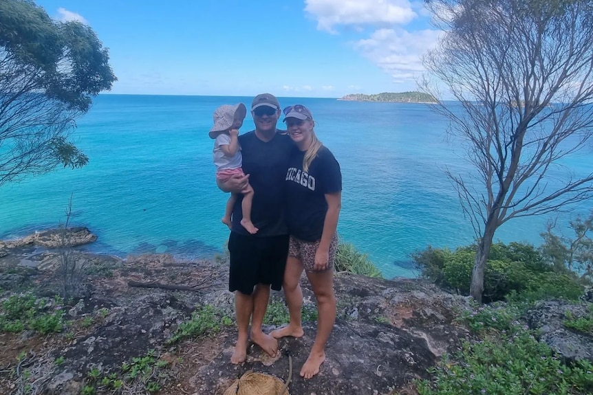 A picture of a man and woman on holiday with their one-year-old, standing in front of blue ocean waters and a clear summer sky. 