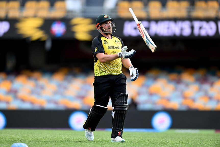 Un hombre vestido con el uniforme de cricket verde y amarillo de Australia lanza suavemente un bate de cricket con frustración.