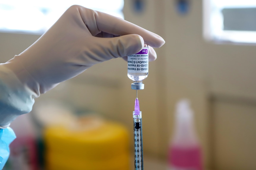 A gloved hand holds a vial above a syringe with the words 'COVID-19 vaccine' written on the label.