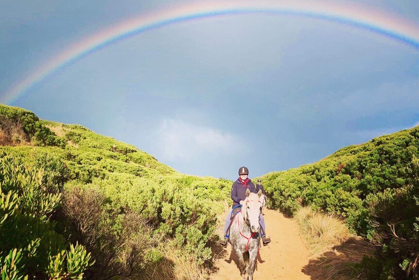A woman rides a horse through a sandy track among scrub, with a rainbow visible overhead.