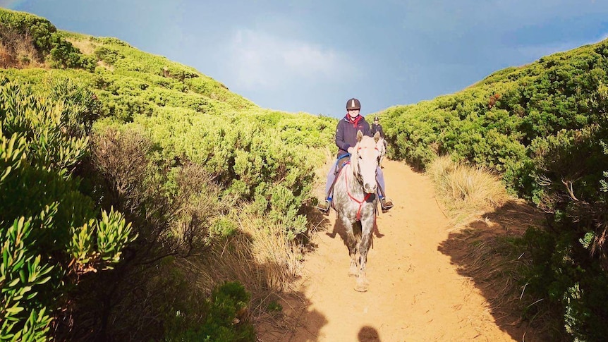 A woman rides a horse through a sandy track among scrub, with a rainbow visible overhead.