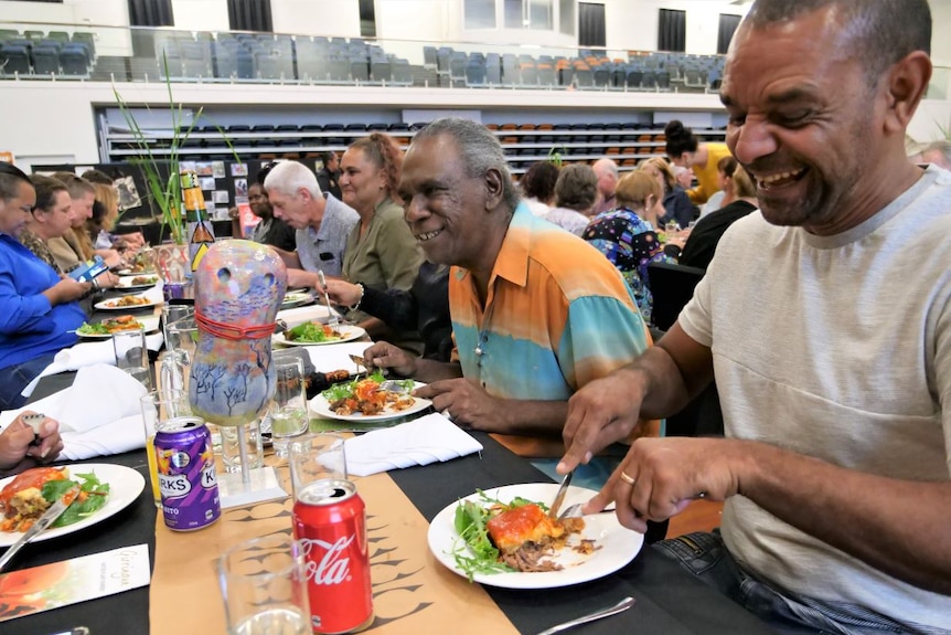 Diners seated at long tables laughing and eating lasagne.