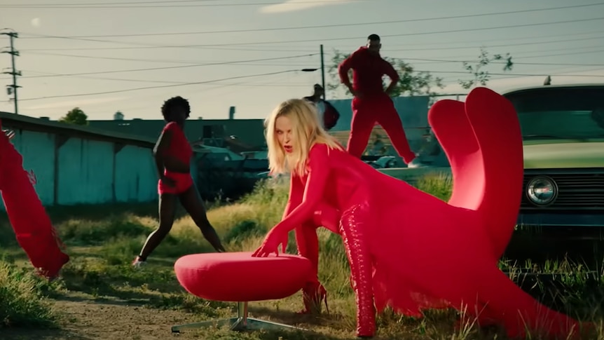 Kylie Minogue sits on a red chair in a yard with dancers. 