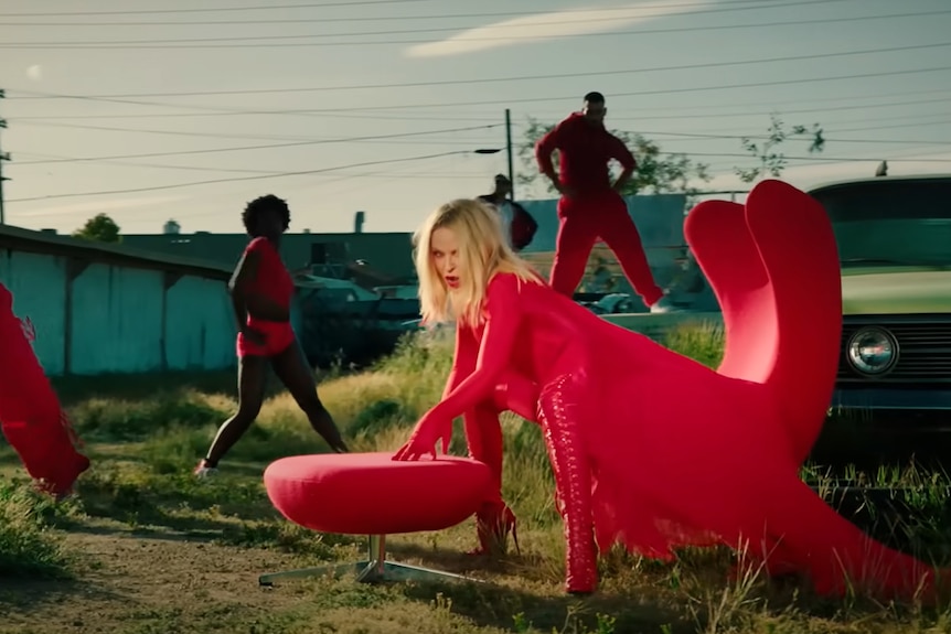 Kylie Minogue sits on a red chair in a yard with dancers. 