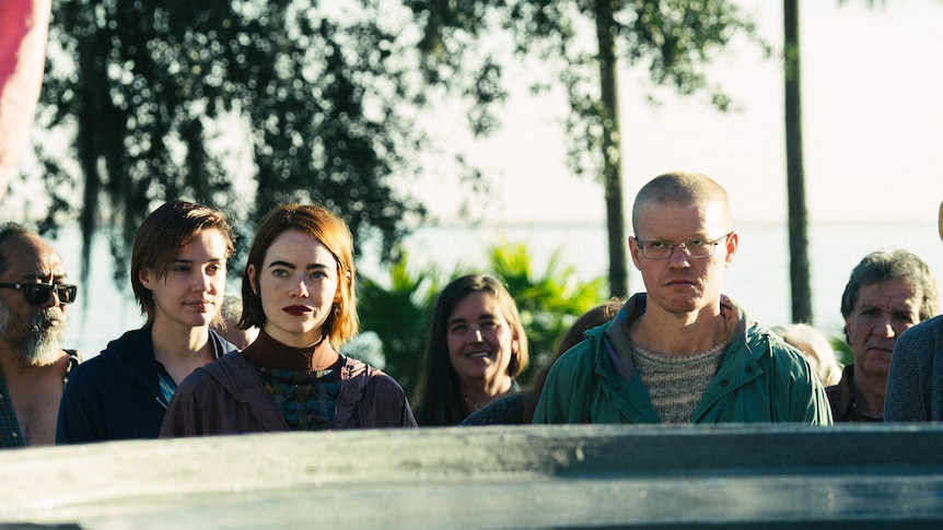 Emma Stone (left) stands next to Jesse Plemons (right) with a few people behind them in front of a casket on a sunny day.
