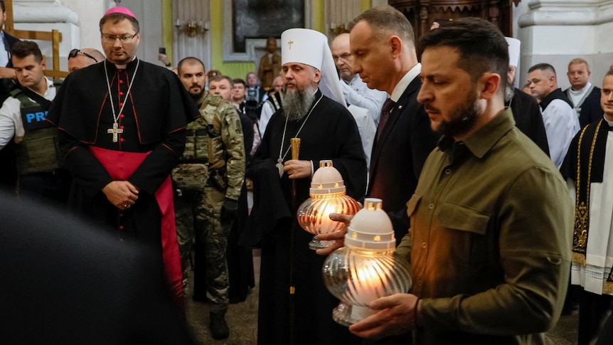 Zelenskyy and another man holding lanterns and looking sorrowful with priests in the background