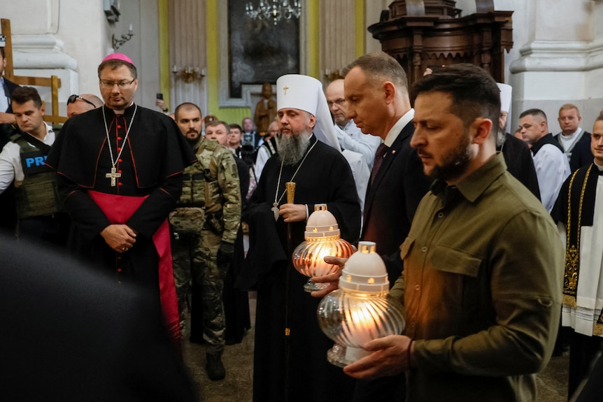 Zelenskyy and another man holding lanterns and looking sorrowful with priests in the background