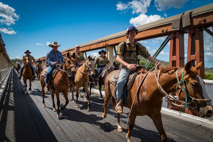 A group of men and women on horseback ride across a wooden truss bridge