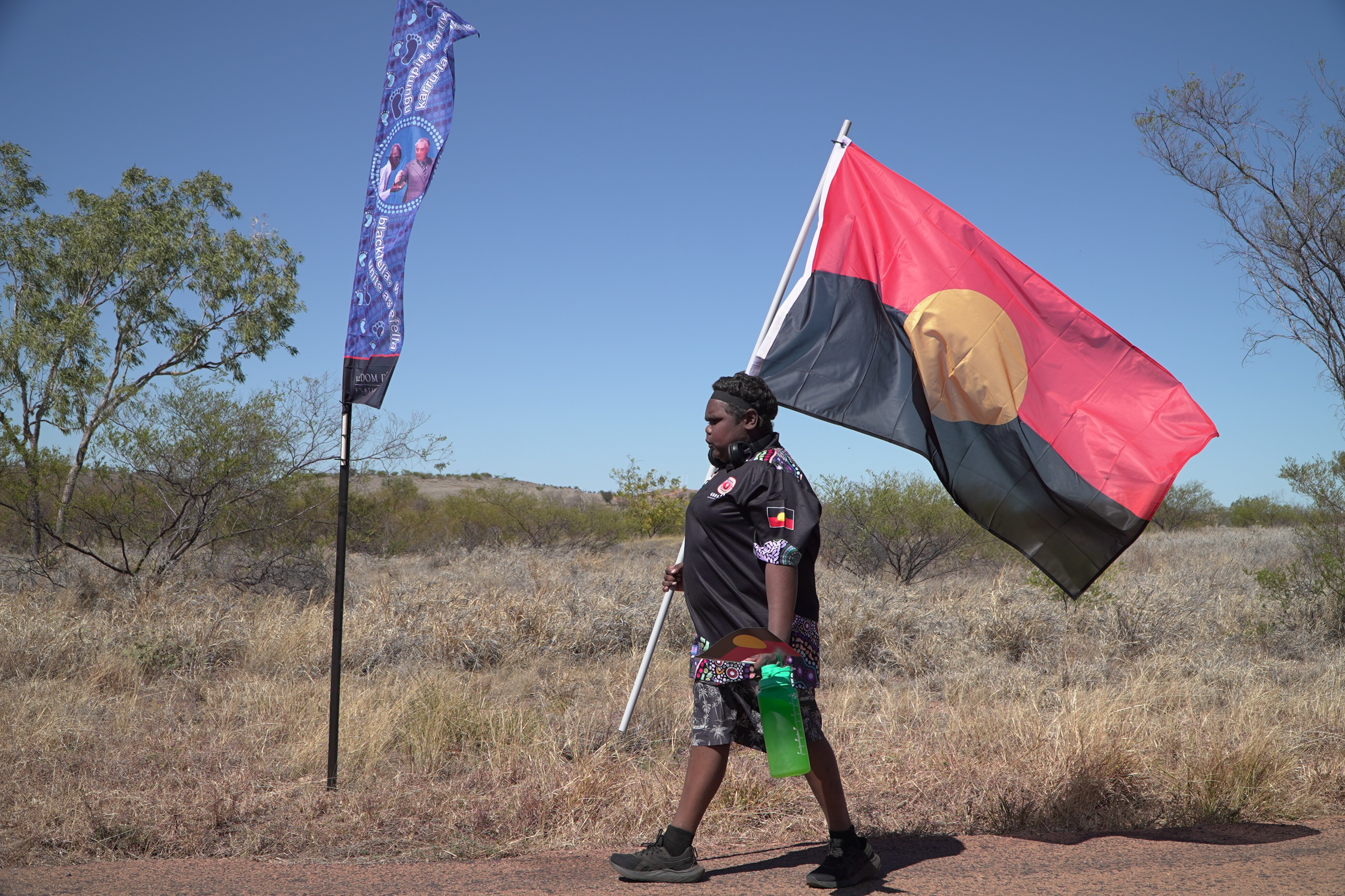 A sole person with dark complexion walking left of frame, holding large Aboriginal flag on pole, outback bush background.