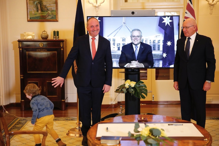 Three men, one on a TV screen video link, stand for a photo, the one on the left grimaces as his young son runs out of frame