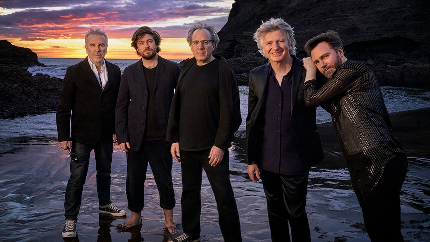 Five members of Crowded House, all in dark suits, standing on a beach at sunset.