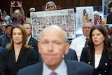 A mans face close up and a women holds a poster with the faces of all those killed in a place crash 