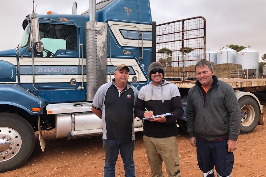 Three farmers standing in front of a semi-trailer being loaded with hay bales. The man in the middle is holding a clipboard