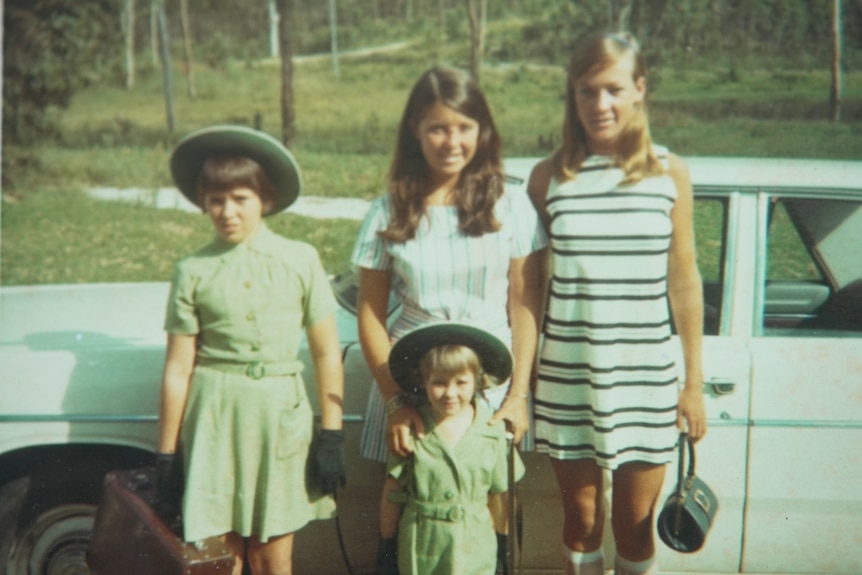 An old photo of three young girls and a young woman standing in front of a car.