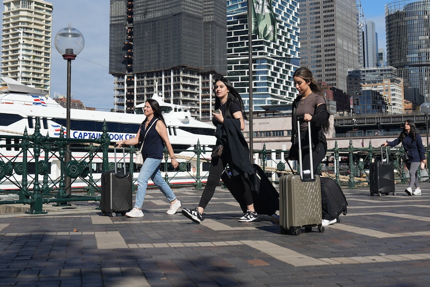 Three young women walk with suitcases at Sydney's Circular Quay