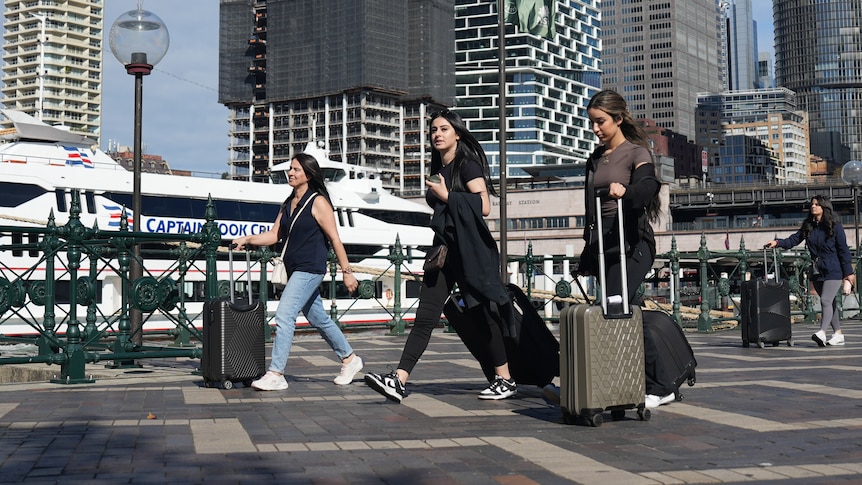 Three young women walk with suitcases at Sydney's Circular Quay