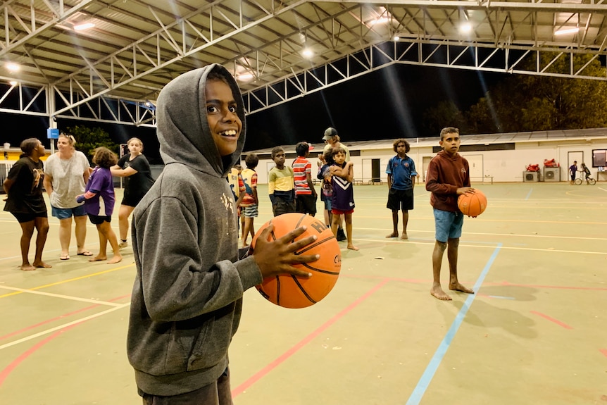 A smiling child prepares to shoot a basketball in a hoop as other kids watch on.
