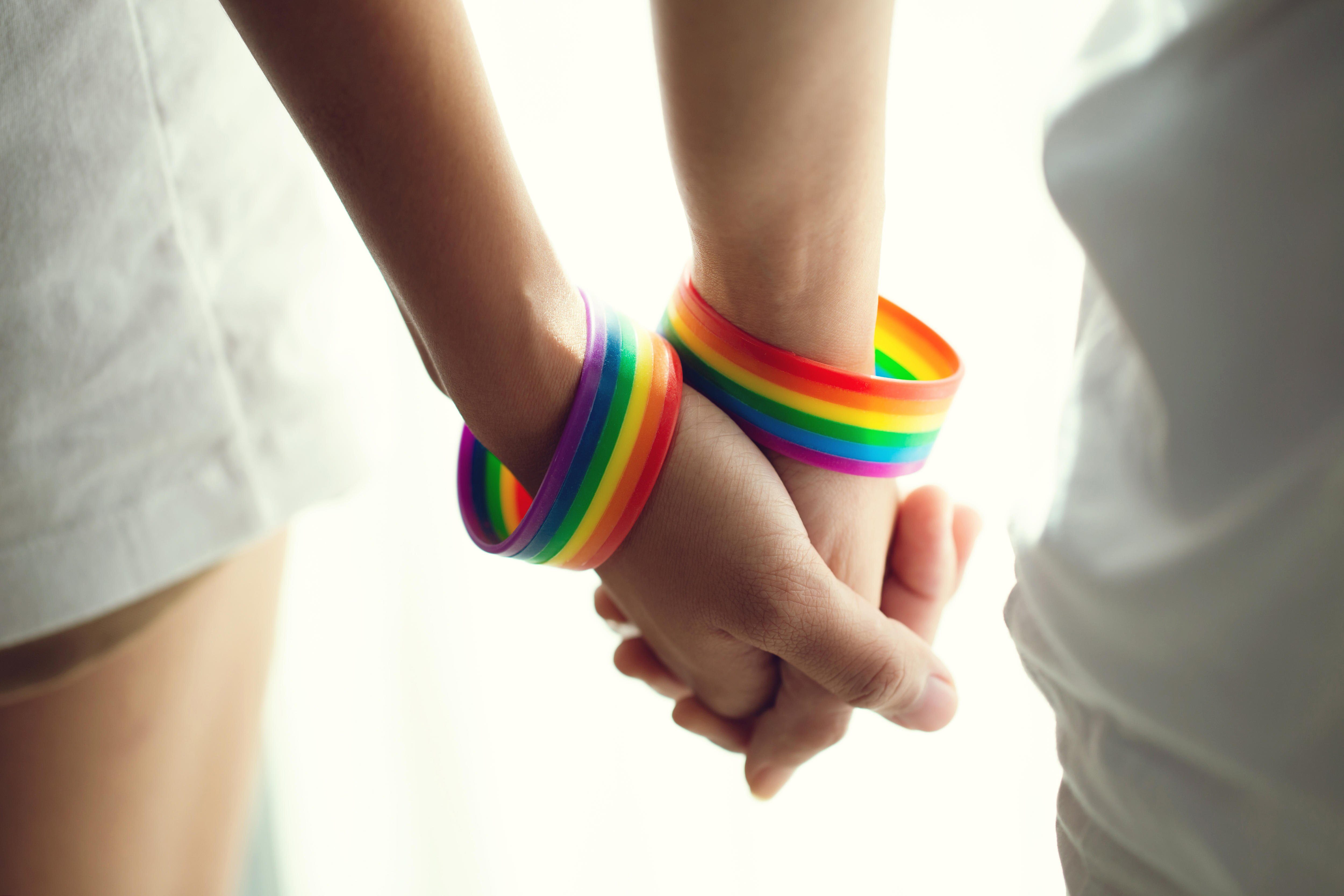 A close-up of two people holding hands while wearing rainbow-coloured wristbands.