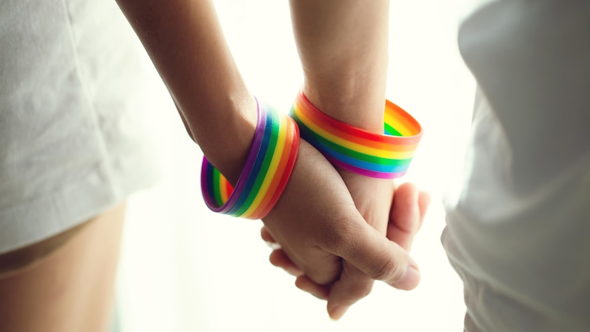 A close-up of two people holding hands while wearing rainbow-coloured wristbands.
