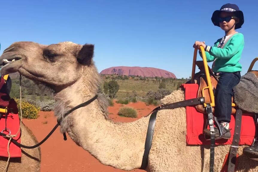Bobby riding a camel with Uluru in the background