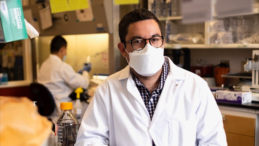 A man wearing glasses, a mask and a lab coat sits at a desk in a lab.