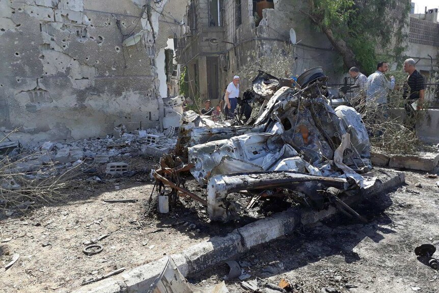 People gather at the scene of an explosion in Damascus. The twisted wreckage of a car is visible.