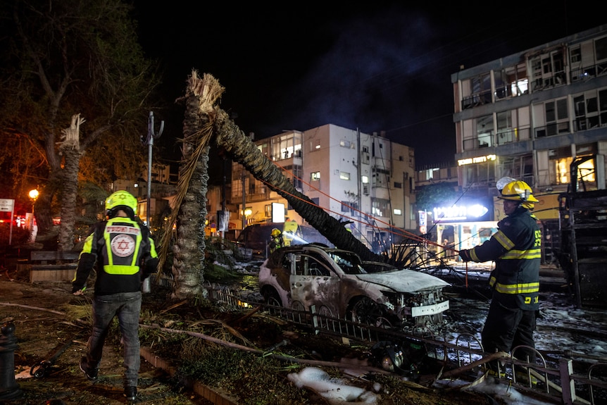 An Israeli firefighter works at an explosion site where a tree has fallen onto a burned out car.