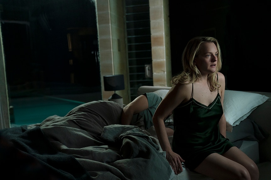 A woman with blonde hair and scared expression sits up in bed in the middle of the night, behind her a man lies asleep.