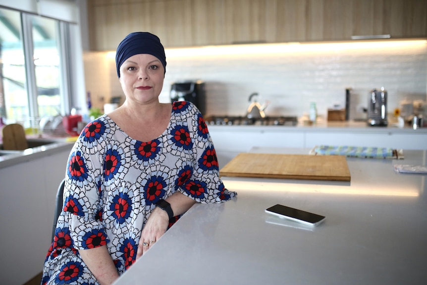A woman wearing a head scarf looking straight at the camera while her mobile phone sits on the kitchen bench top