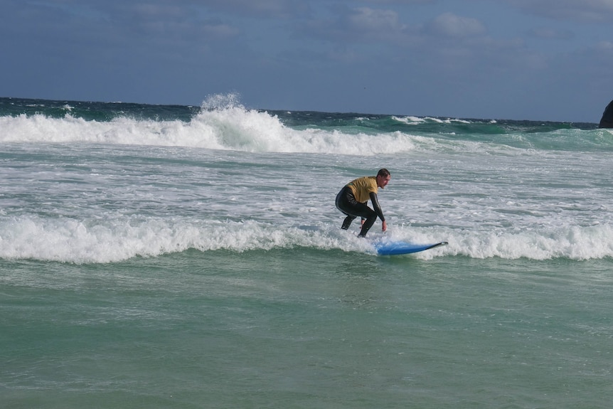 A man in a wetsuit and yellow rashie standing on a surfboard in the whitewash at a beach