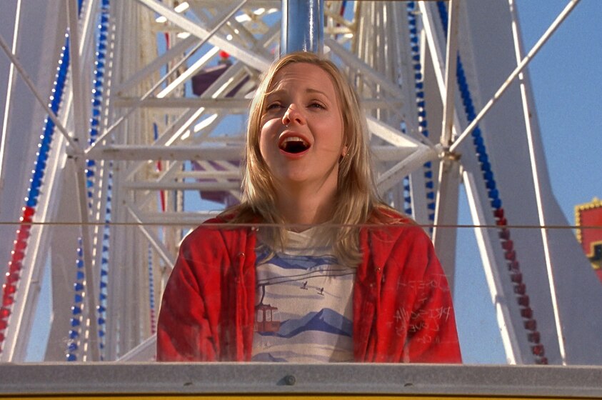 A blonde woman in red cardigan sits alone on ferris wheel ride on clear day with stoned or dazed expression.