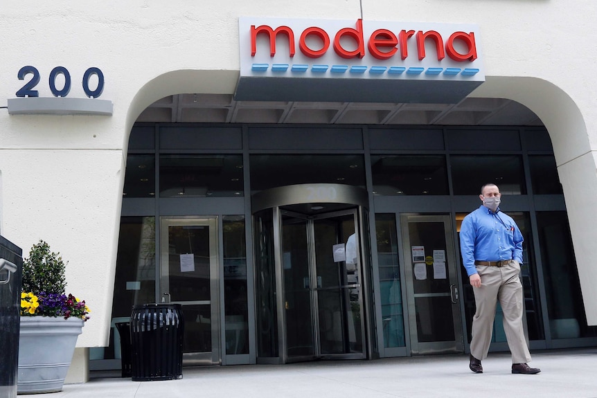 A man in a face mask walks outside the glass doors of Moderna Inc's offices, with Moderna's logo mounted above