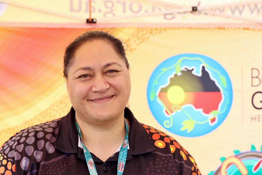 A woman with brown hair pulled back in a bun smiles, standing in front of a banner with the Aboriginal flag.