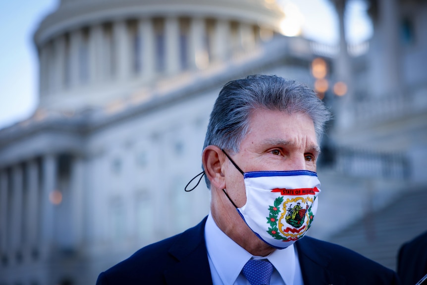 A man in a suit and a face mask bearing the West Virginia state flag stands in front of the US Capitol building