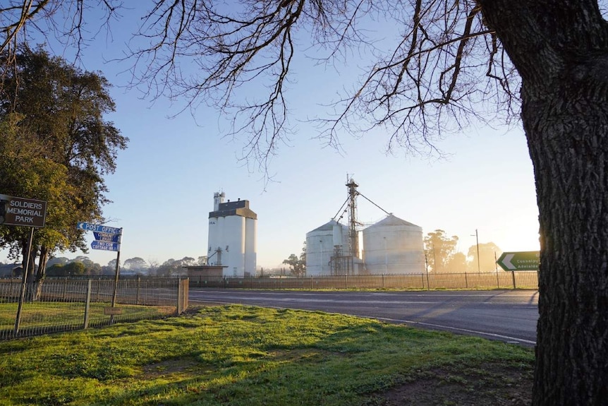 An early sunrise shines through two large silos. A big tree and sign post reading post office and cottage in the foreground.