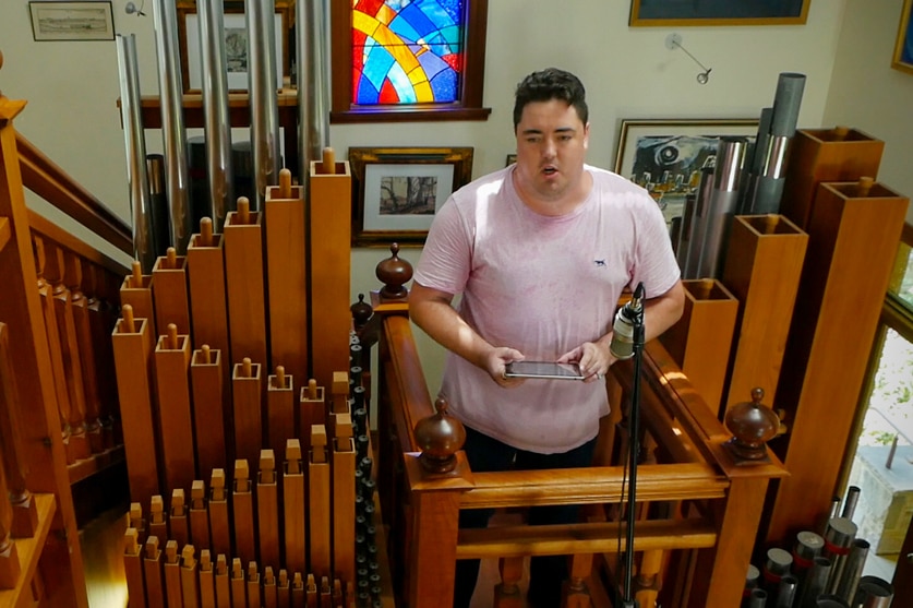 Man on a small wooden balcony standing within the top of a pipe organ surrounded by wooden and metal pipes.