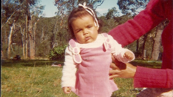 A small rolly-polly child is being held up by mother's arms. Behind is bright green Australian bushland.