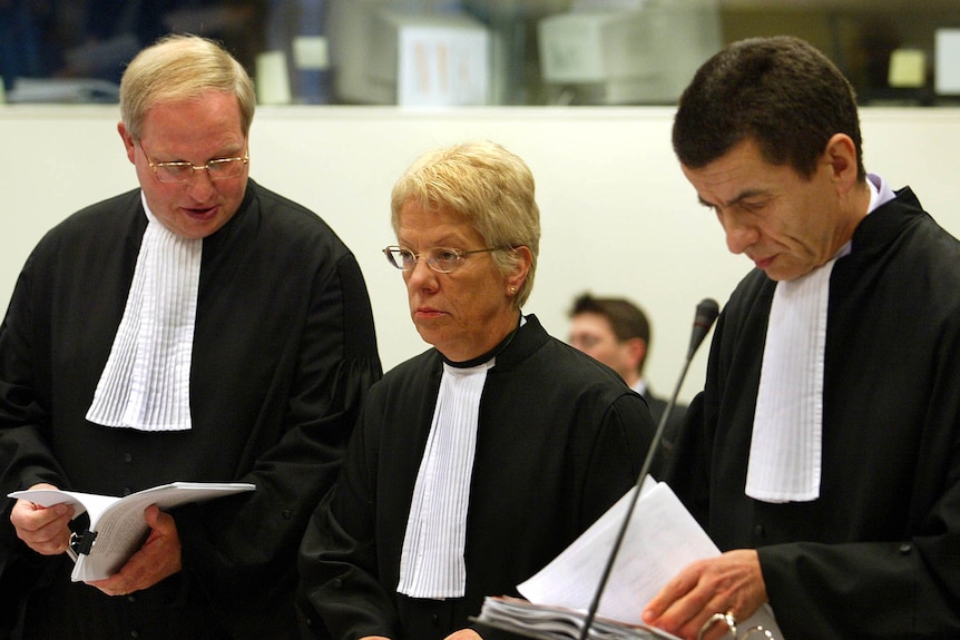 Two men and a woman in black barrister robes with white jabots standing at a microphone 