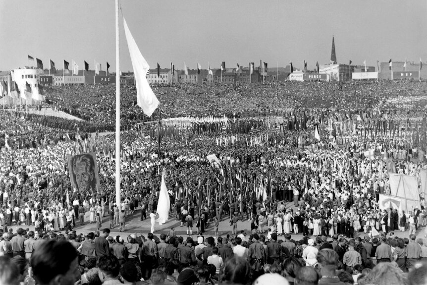 Black and white photo of thousands of young people in a flag adorned stadium. They're grouped in sections and many carry flags.
