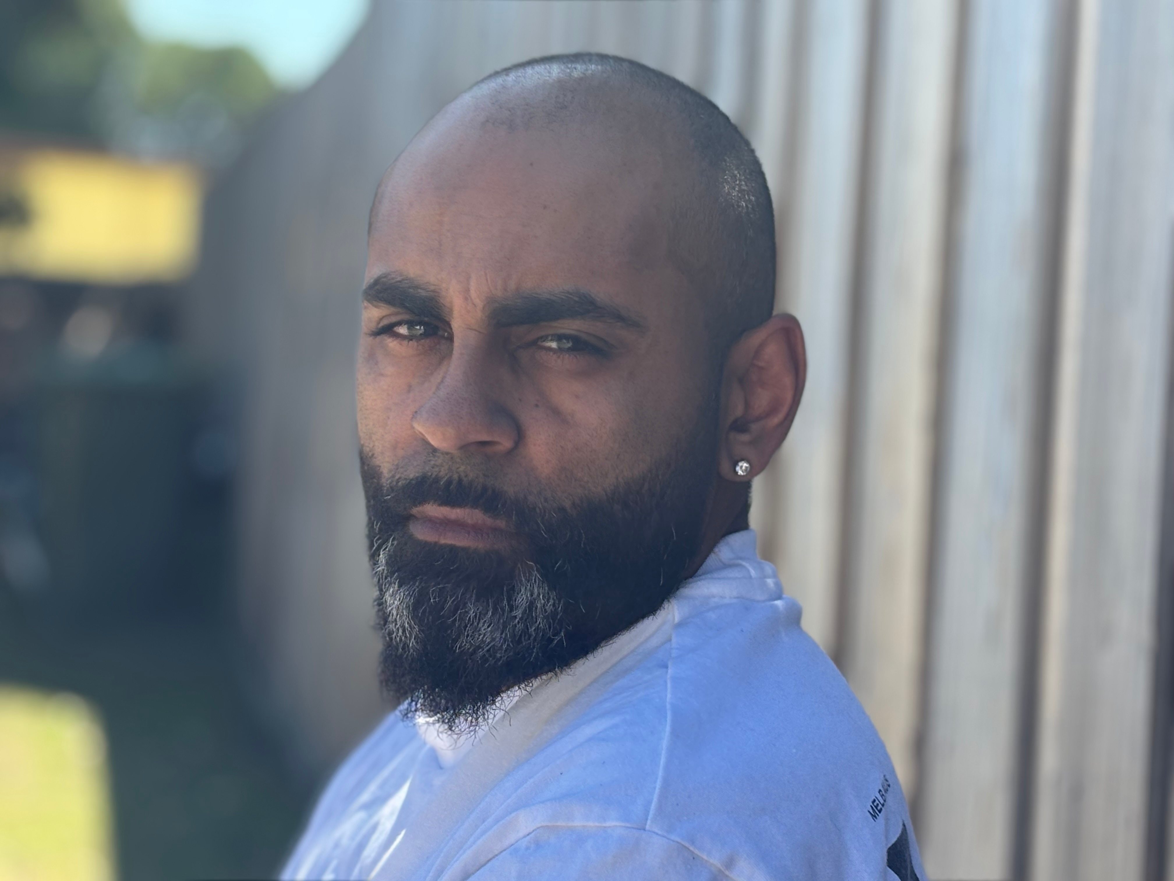 Um homem indígena com barba perto de uma cerca de madeira. 