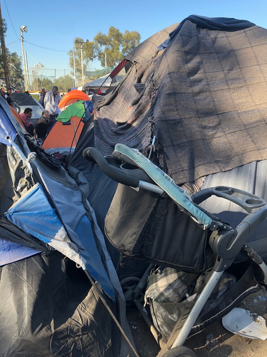 A pram sits near a line of tents covered with cloth while people sit among their makeshift accommodations.