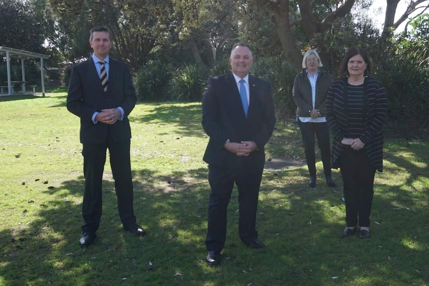 Wide shot of a group of people including two men in suits and two women standing on grass.