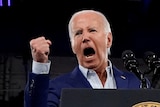 Joe Biden yells mouth wide open with his fist in the air at a podium.
